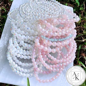 Op de afbeelding: Een verzameling kralenarmbanden in heldere, witte en roze tinten. De armbanden zijn gemaakt van kleine, ronde kralen en zijn opgestapeld. Het websiteadres "www.angelic-kisses.com" is zichtbaar.