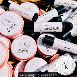 Peut inclure: Plusieurs baumes &agrave; l&egrave;vres Angelip biologiques, pr&eacute;sent&eacute;s dans des bo&icirc;tes roses et des tubes blancs. Les baumes &agrave; l&egrave;vres ont des bouchons noirs et affichent le nom et le logo de la marque. Le site web "www.angelic-kisses.com" est visible en bas.