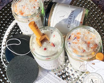 May include: An assortment of glass jars filled with bath salts, some with wooden scoops. The jars are labeled with words like "Creator" and "Serenity." The salts are white, with colorful flower petals. The image includes a website address.