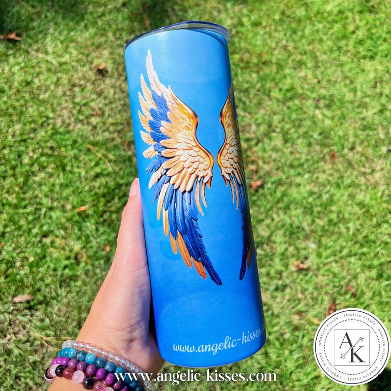 May include: A blue tumbler featuring a design of angel wings in shades of gold, blue, and orange. The tumbler has a silver lid and the website address www.angelic-kisses.com is printed on the bottom. The tumbler is held against a background of green grass.
