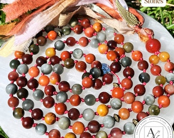 May include: A long beaded necklace featuring round, polished stones in shades of red, orange, brown, and grey, strung on a red cord with knotted spacers. The necklace includes a bronze-colored wing charm and frayed fabric ribbons.
