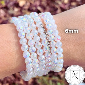 Op de afbeelding: Een stapel iriserende witte kralenarmbanden. De kralen zijn 6 mm groot en hebben een glinsterende, opalescente kwaliteit. De armbanden worden op een pols getoond, met het websiteadres "www.angelic-kisses.com" zichtbaar.