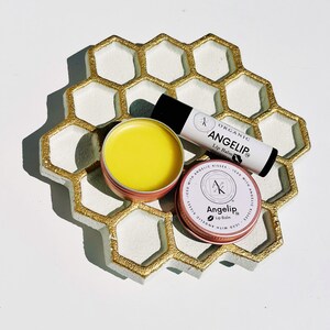 Peut inclure: Un plateau en forme de nid d'abeille avec des bordures dor&eacute;es contient trois produits de baume &agrave; l&egrave;vres. Il y a un tube avec le texte "ANGELIP Lip Balm" et deux petites bo&icirc;tes rondes. Une bo&icirc;te est ouverte, r&eacute;v&eacute;lant un baume jaune. L'autre bo&icirc;te porte &eacute;galement le texte "ANGELIP Lip Balm".