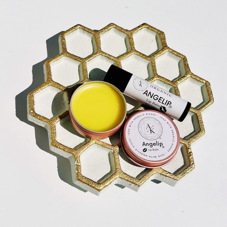 Peut inclure: Un plateau en forme de nid d'abeille avec des bordures dor&eacute;es contient trois produits de baume &agrave; l&egrave;vres. Il y a un tube avec le texte "ANGELIP Lip Balm" et deux petites bo&icirc;tes rondes. Une bo&icirc;te est ouverte, r&eacute;v&eacute;lant un baume jaune. L'autre bo&icirc;te porte &eacute;galement le texte "ANGELIP Lip Balm".