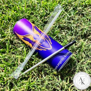 May include: A blue and purple tumbler with a gold wing design, a clear plastic straw, and a silver metal straw with a black tip. The tumbler has the website address www.angelic-kisses.com printed on it. The items are on a bed of green grass.