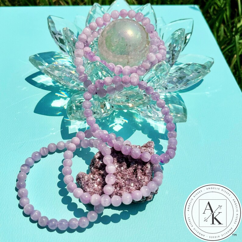 May include: Three light purple beaded bracelets are draped over a clear crystal lotus flower and a small rock. The bracelets are made of small, round beads. The background is a light blue surface with a shadow.