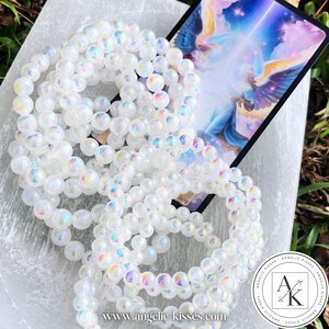 Op de afbeelding: Verschillende iriserende witte kralenarmbanden zijn gerangschikt op een wit oppervlak, met een kleurrijke illustratiekaart op de achtergrond. De armbanden zijn gemaakt van ronde, gepolijste kralen die een regenboog van kleuren reflecteren. De afbeelding bevat ook een logo en websiteadres.