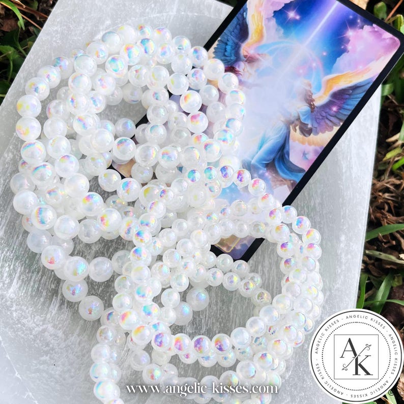 Op de afbeelding: Verschillende iriserende witte kralenarmbanden zijn gerangschikt op een wit oppervlak, met een kleurrijke illustratiekaart op de achtergrond. De armbanden zijn gemaakt van ronde, gepolijste kralen die een regenboog van kleuren reflecteren. De afbeelding bevat ook een logo en websiteadres.