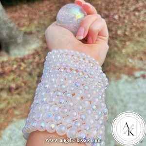 Op de afbeelding: Een stapel iriserende, heldere kralenarmbanden siert een pols, met een bijpassende bol in de hand. De kralen reflecteren een regenboog aan kleuren. Het websiteadres www.angelickisses.com is zichtbaar.