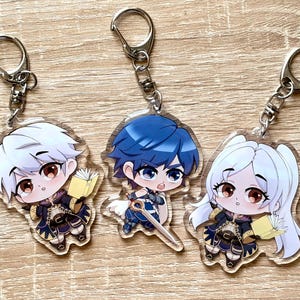 Puede incluir: Tres llaveros de acrílico con personajes de anime en estilo chibi. El personaje de la izquierda tiene el pelo blanco y sostiene un libro. El del centro tiene el pelo azul y sostiene una espada. El de la derecha tiene el pelo blanco largo y sostiene un libro.