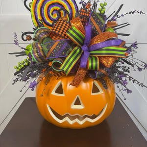 May include: A Halloween-themed centerpiece featuring an orange jack-o'-lantern with a carved face. The pumpkin is adorned with a large, colorful lollipop, ribbons, and decorative accents in orange, purple, green, and black. The arrangement sits on a dark brown surface.