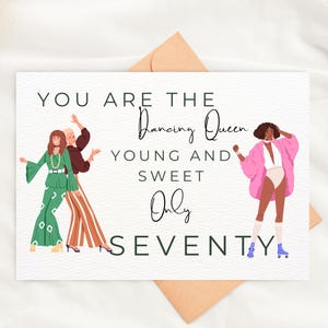 Puede incluir: Una tarjeta de felicitación blanca con el texto "YOU ARE THE Dancing Queen YOUNG AND SWEET Only SEVENTY." La tarjeta presenta ilustraciones de tres mujeres con ropa de los años 70, incluida una mujer con patines. Un sobre marrón está detrás de la tarjeta.