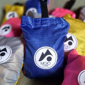 Puede incluir: Una bolsa de compras reutilizable azul con un mosquetón negro y un logotipo blanco que dice "MOJO CLIMB". La bolsa está rodeada de otras bolsas de colores.