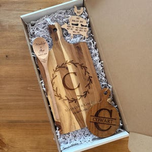 Puede incluir: Un juego de regalo de madera en una caja, que incluye una tabla de cortar con una inicial grande y un diseño de corona de laurel, una cuchara de madera y una pequeña tabla de cortar redonda. El juego está personalizado con nombres y una fecha.