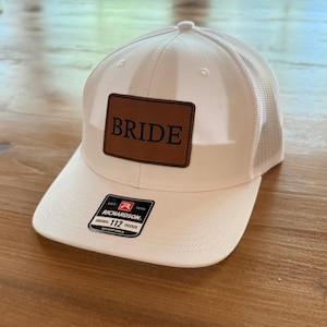 Peut inclure: Casquette de camionneur crème avec un dos en filet et une visière incurvée. Le chapeau comporte un patch en cuir marron avec le mot "BRIDE" en lettres noires. Un autocollant noir et blanc est sur la visière.