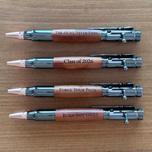 Pode incluir: Quatro canetas com um design em forma de bala. As canetas têm um esquema de cores prateado e castanho, com texto que diz "The Hunt Never Ends", "Class of 2026", "Forge Your Path" e "Built Not Given".