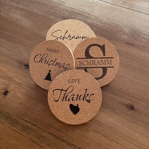 Puede incluir: Cuatro posavasos de corcho con diferentes diseños. Un posavasos dice "Feliz Navidad" con un pequeño árbol de Navidad. Otro posavasos dice "Dar gracias" con un pavo. Un tercer posavasos dice "Schramm" con una gran "S" y el nombre "Schramm" debajo. El último posavasos dice "Schramm" en cursiva.