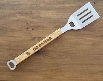 May include: A stainless steel spatula with a wooden handle. The handle is engraved with the words "GO DAWGS" and a small logo. The spatula has a bottle opener on the end of the handle.