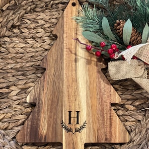 Può includere: Un tagliere di legno a forma di albero di Natale con una finitura in legno naturale. Il tagliere ha un foro nella parte superiore per appenderlo ed è inciso con la lettera "H" circondata da una ghirlanda.