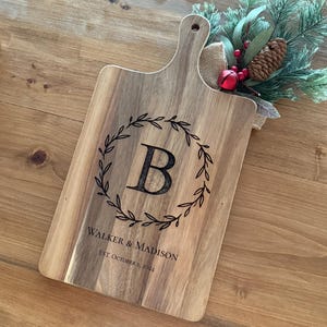 Könnte beinhalten: Ein Schneidebrett aus Holz mit Griff, das mit einem Monogramm "B" in einem Kranzdesign verziert ist. Das Brett ist mit dem Text "Walker & Madison Est. October 1, 2024" graviert.