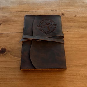 May include: A dark brown leather journal with a wrap-around strap. The cover is embossed with the words "MAN OF GOD" and a cross, along with the text "NO WEAPON FORMED... AGAINST YOU SHALL PROSPER."