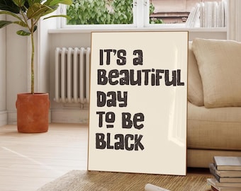 May include: A framed poster with the text "It's a Beautiful Day To Be Black" in a bold, black font. The poster is displayed indoors, leaning against a wall near a window, with a plant in a terracotta pot visible.