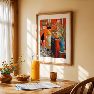 K&ouml;nnte beinhalten: Ein gerahmter Kunstdruck zeigt eine Frau in einem gelben Kleid, die in einer roten K&uuml;che mit einer schwarzen Katze kocht. Eine Blumenvase, eine Glasflasche Saft und ein Holztisch befinden sich im Vordergrund.