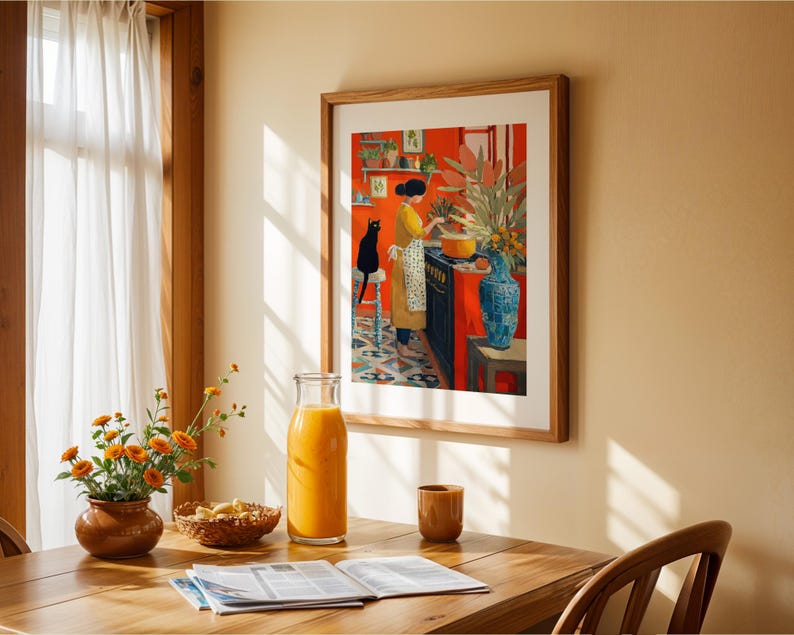 K&ouml;nnte beinhalten: Ein gerahmter Kunstdruck zeigt eine Frau in einem gelben Kleid, die in einer roten K&uuml;che mit einer schwarzen Katze kocht. Eine Blumenvase, eine Glasflasche Saft und ein Holztisch befinden sich im Vordergrund.