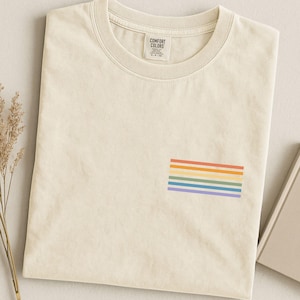 May include: A cream-colored crew neck t-shirt featuring a small graphic of six horizontal stripes in orange, yellow, light green, light blue, and purple on the lower right side. The inner neck label displays the text Comfort Colors.