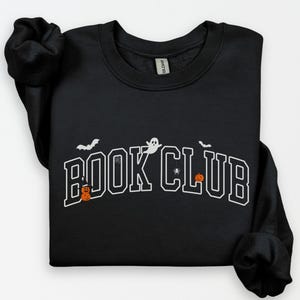 May include: Black sweatshirt with the text "BOOK CLUB" in white, arched across the front. The design includes a ghost, bats, a spider, and jack-o'-lanterns, all in a Halloween theme.