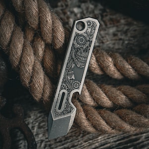 May include: A small, silver pocket knife with a detailed engraved design featuring a skull and mechanical elements. The knife has a bottle opener and a pointed blade. The background includes a thick, brown rope.