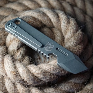 May include: A small, gray pocket knife with a clip and a partially serrated edge. The knife features a geometric design and a small emblem. It rests on a thick, textured rope.