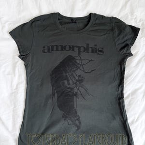 May include: Dark gray t-shirt featuring the band name "amorphis" in black block letters above a black graphic of a figure with long hair. The text "YESTERDAY'S PLAYGROUND" is printed in green at the bottom of the shirt.