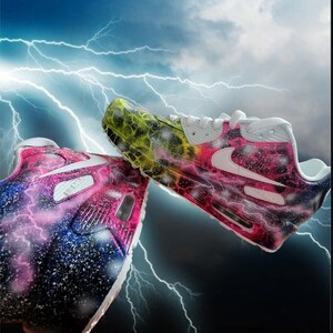 May include: A pair of white trainers with a galaxy-themed paint design. One shoe features a pink, blue, and white galaxy design with white lightning bolts. The other shoe features a yellow, green, and black galaxy design with white lightning bolts.