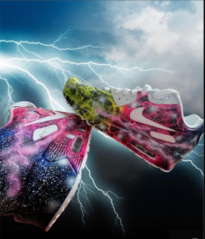 May include: A pair of white trainers with a galaxy-themed paint design. One shoe features a pink, blue, and white galaxy design with white lightning bolts. The other shoe features a yellow, green, and black galaxy design with white lightning bolts.