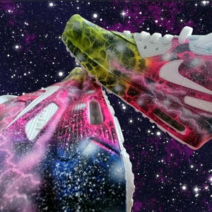 May include: A pair of white trainers with a galaxy-themed paint design. The trainers have pink, blue, and yellow accents. The design features a swirling galaxy pattern with stars and lightning bolts.