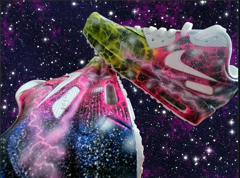 May include: A pair of white trainers with a galaxy-themed paint design. The trainers have pink, blue, and yellow accents. The design features a swirling galaxy pattern with stars and lightning bolts.