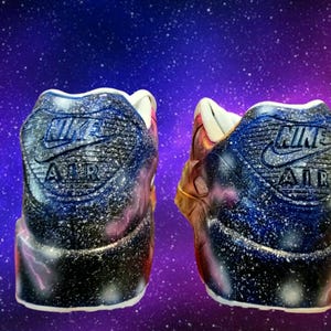 May include: A pair of custom-painted Nike Air Max 90 trainers with a galaxy print. The trainers are black, blue, and white with a pink and gold accent. The Nike Air logo is visible on the back of each shoe.
