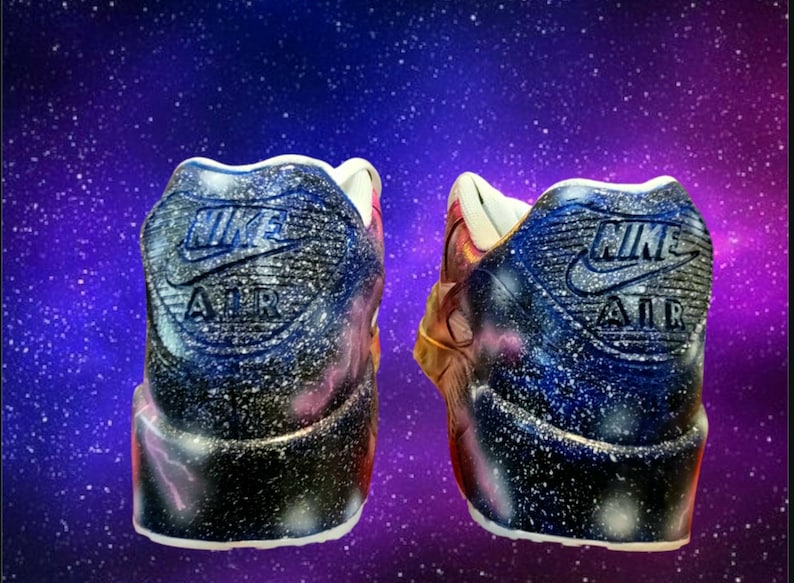 May include: A pair of custom-painted Nike Air Max 90 trainers with a galaxy print. The trainers are black, blue, and white with a pink and gold accent. The Nike Air logo is visible on the back of each shoe.