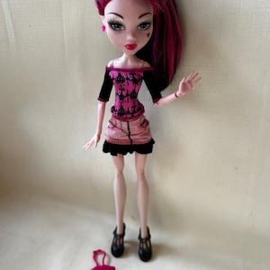 May include: A doll with black and pink hair, wearing a pink and black outfit. The doll has pale skin, red lips, and heart-shaped facial details. Black shoes and a pink purse are at the doll's feet. The doll is a collectible toy.