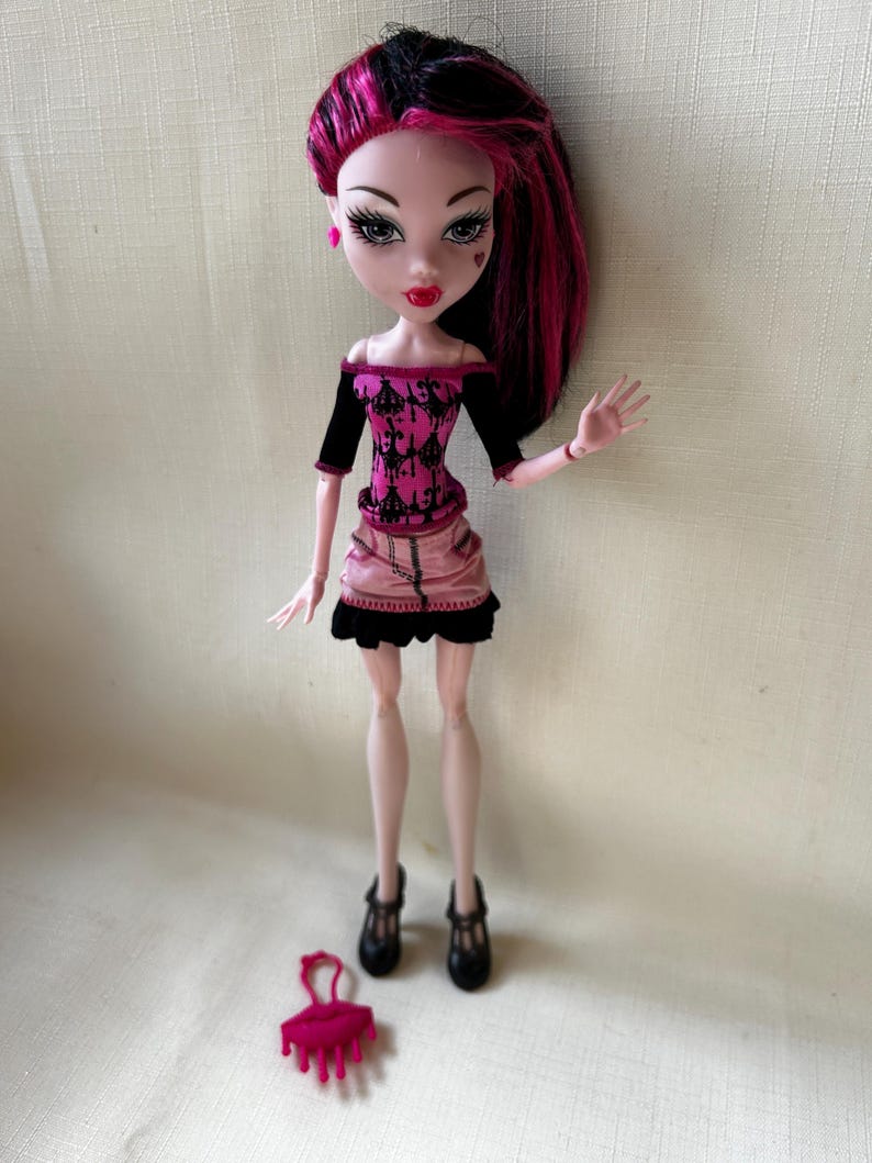 May include: A doll with black and pink hair, wearing a pink and black outfit. The doll has pale skin, red lips, and heart-shaped facial details. Black shoes and a pink purse are at the doll's feet. The doll is a collectible toy.