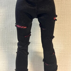 Puede incluir: Pantalones negros para mu&ntilde;eca con detalles desgastados. Los pantalones tienen varios rasgu&ntilde;os y roturas, revelando una tela roja debajo. Los pantalones son de un material texturizado.