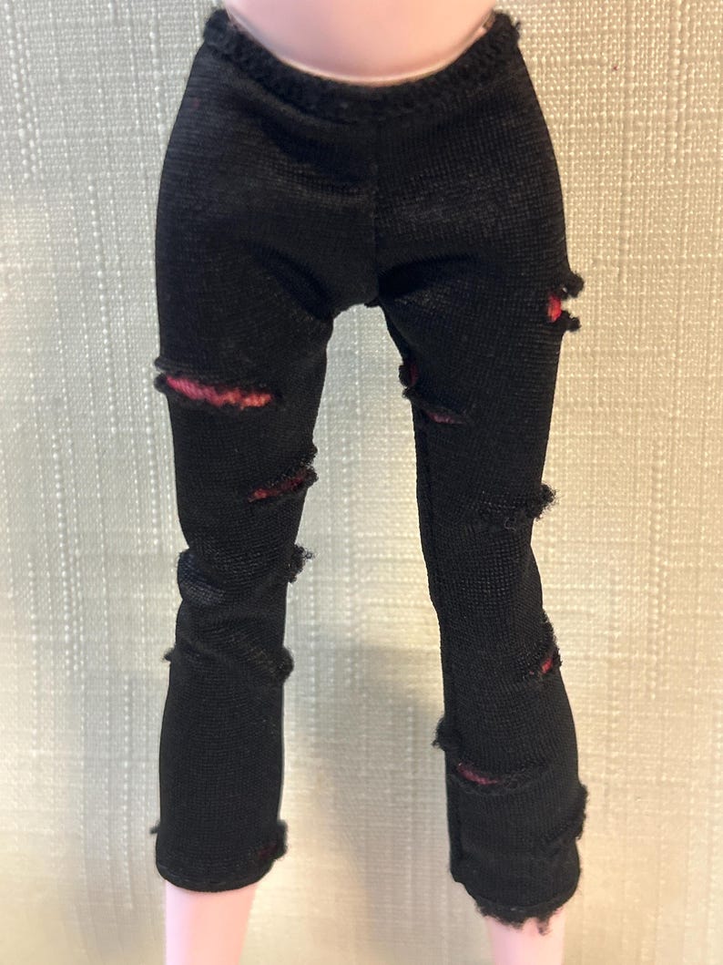 Puede incluir: Pantalones negros para mu&ntilde;eca con detalles desgastados. Los pantalones tienen varios rasgu&ntilde;os y roturas, revelando una tela roja debajo. Los pantalones son de un material texturizado.