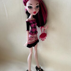 May include: A doll with black and pink hair, wearing a pink and black top and skirt, and black heels. The doll is holding a red purse. The doll has pale skin, red lipstick, and heart-shaped details on its face.
