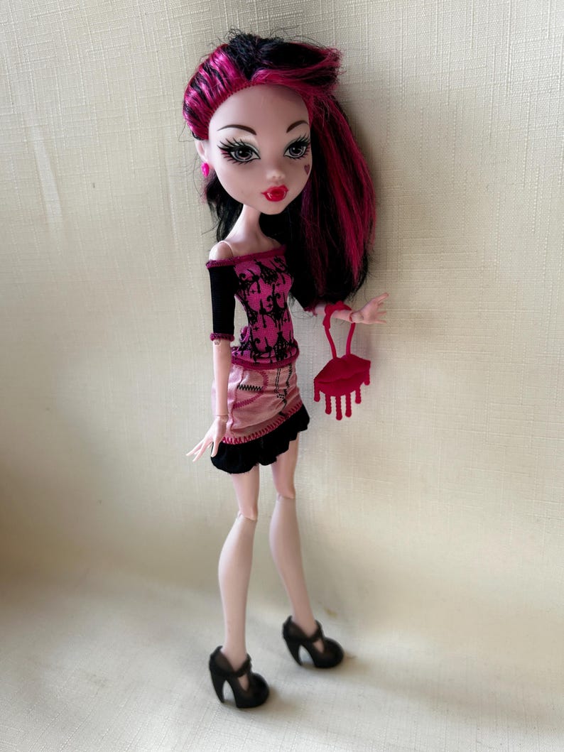 May include: A doll with black and pink hair, wearing a pink and black top and skirt, and black heels. The doll is holding a red purse. The doll has pale skin, red lipstick, and heart-shaped details on its face.