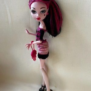 May include: A fashion doll with pale skin, red lips, and heart-shaped face details. The doll has black and pink hair, wears a pink and black outfit, and black high heels. A small pink purse is held in the doll's hand.