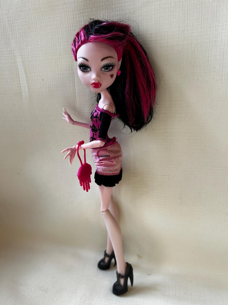 May include: A fashion doll with pale skin, red lips, and heart-shaped face details. The doll has black and pink hair, wears a pink and black outfit, and black high heels. A small pink purse is held in the doll's hand.