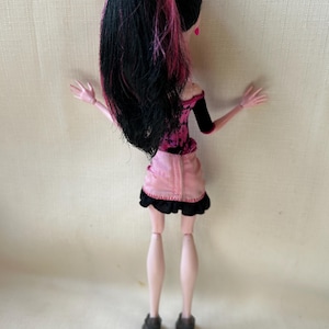 May include: A doll with black hair streaked with pink, styled in a high ponytail. The doll wears a pink and black outfit, including a short skirt with a black trim. The doll's legs are pale pink, and it wears black high-heeled shoes.