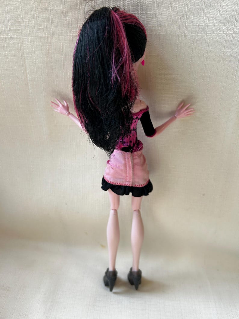 May include: A doll with black hair streaked with pink, styled in a high ponytail. The doll wears a pink and black outfit, including a short skirt with a black trim. The doll's legs are pale pink, and it wears black high-heeled shoes.