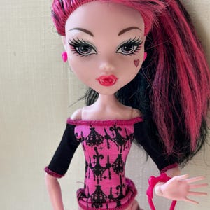 May include: A doll with pink and black hair, wearing a pink and black top and a pink skirt. The doll has pink earrings, a heart-shaped mark on its cheek, and is holding a pink purse. The doll's lips are red.